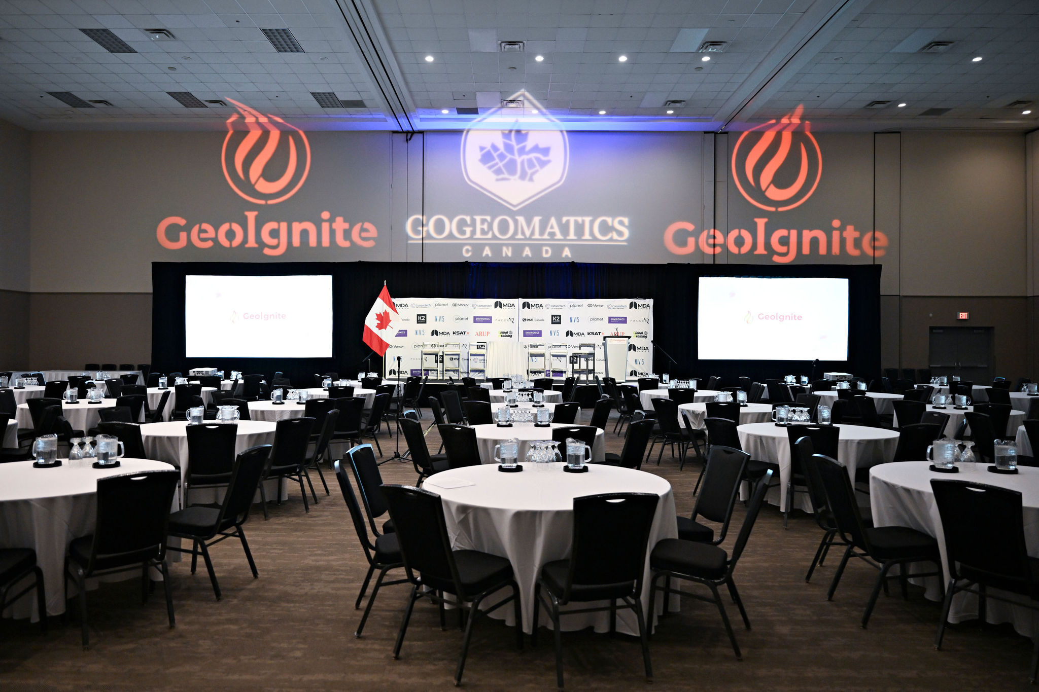 The GeoIgnite plenary ballroom prepared with round tables, stage branding, sponsor wall and a Canadian flag.